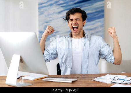 Excited happy successful Hispanic guy, male freelancer, in casual clothes, sits at workplace, rejoicing in victory, gain or big profit, gesturing with his hands, shouting emotionally Stock Photo