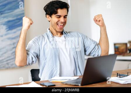Young excited joyful hispanic man, manager or freelancer, in casual clothes, sitting at workplace with laptop, happy with victory or big profit, make deal, gesturing with his hands, smiling happily Stock Photo
