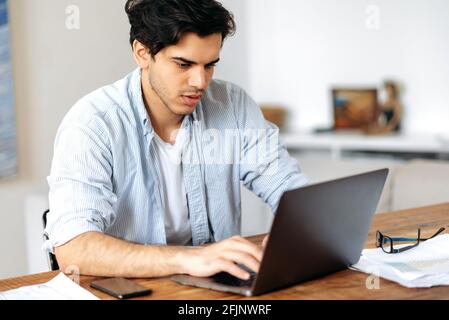 Young serious successful mixed race man, freelancer, manager or student, working or studying remotely, in casual wear, uses laptop, texting with colleague or client, browsing internet, searching ideas Stock Photo