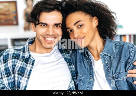 Joint photo of a hispanic guy with african american girl. Close-up portrait of happy young mixed race family couple sitting in living room, wearing casual stylish clothes, looking at camera, smiling Stock Photo
