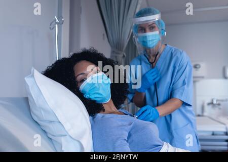 Caucasian female doctor in hospital in face mask examining female patient with stethoscope Stock Photo