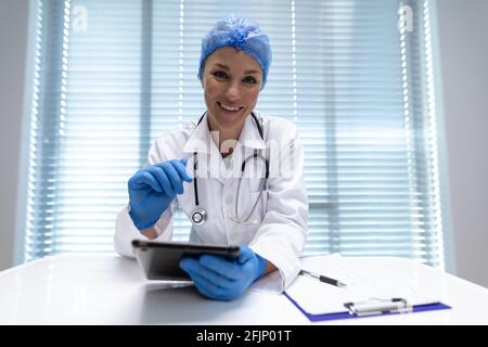 Caucasian female doctor at desk using tablet and smiling during video call consultation