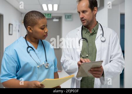 Couple of diverse male and female doctors walking through corridor looking at tablet Stock Photo