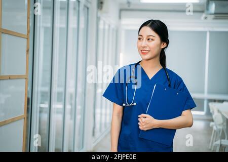 Young beautiful Asian female doctor in uniform with stethoscope standing in hospital hallway holding clipboard checking reports while looking away Stock Photo