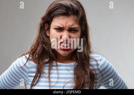 Angry young woman gnashing her teeth and glaring at the camera with a furious expression in a head and shoulders portrait on grey Stock Photo