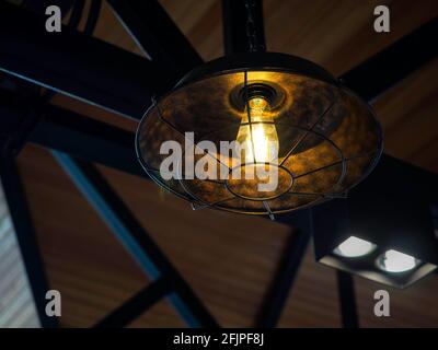 Vintage metal frame pendant lamp. Close-up of shining electric ceiling light with bulb decoration hanging from the ceiling in the dark. Stock Photo