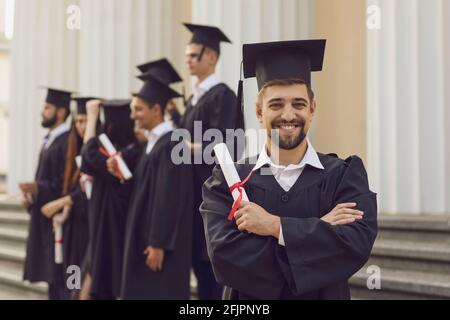 Man in graduation gowns holding diploma and smiling while his friends standing in the background Stock Photo