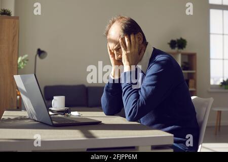 Middle aged man freelancer holding head with hands sitting front of laptop Stock Photo
