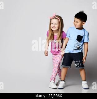Little cute frolic kids Asian boy and blonde girl play hide and seek together, stand holding hands looking at copy space Stock Photo