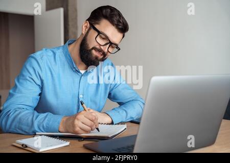 Millennial male student making notes watching webinar on laptop computer Stock Photo