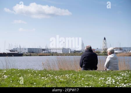 Older retired couple sits the daisy-covered spring grass on the edge of the IJsselmeer on a sunny day with an industrial cityscape of Lemmer Stock Photo