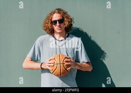 horizontal portrait of a thin long-haired caucasian young man . He is holding a basketball ball with both hands. Looking at camera serious Stock Photo