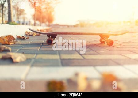 Skateboarding. Skateboard stands on the road, next to the fallen leaves. Side view. In the background, an empty street of the city. Sunlight. Copy spa Stock Photo
