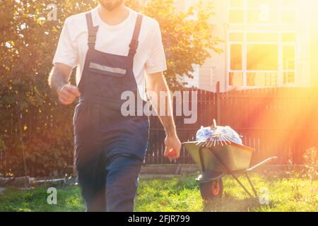 close-up of a man in a blue uniform at sunset ,in motion,the gardener finished the work .cart with bags and rake in the background. Stock Photo