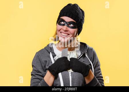A female hacker in a black hat and a mask-glasses, greedily holds a wad of money in her hands and smiles. Yellow background. The concept of crime and Stock Photo