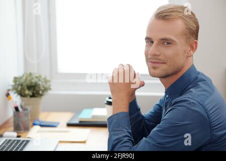 Portrait of handsome young blond businessman sitting at office desk and smiling at camera after finishing work on big project Stock Photo
