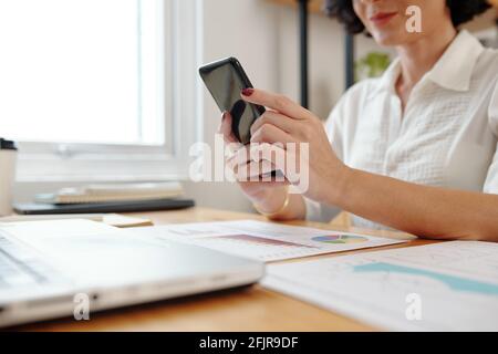 Hands of female entrepreneur sitting at working table with various documents and answering text messages on her phone Stock Photo