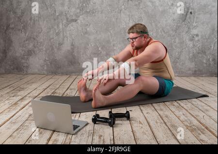 Funny fat man in sportswear doing sports exercises looking into a laptop. home workout  Stock Photo