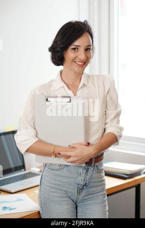 Portrait of smiling young female entrepreneur with clipboard in hands looking at camera Stock Photo