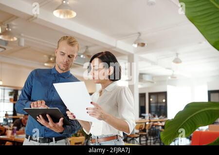 Serious young business partners standing in office cafe and discussing important data in report Stock Photo