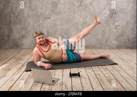funny fat man in sports clothes doing exercises. He plays sports remotely while looking at his laptop Stock Photo