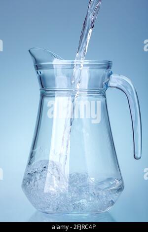 glass transparent jug is filled with a stream of water splashes at the bottom of the container, kitchen utensils isolated on a light cold background. Stock Photo