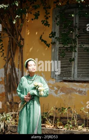 Atractive young Asian woman in traditional dress standing outdoors, closing eyes and enjoying summer rays of sun Stock Photo