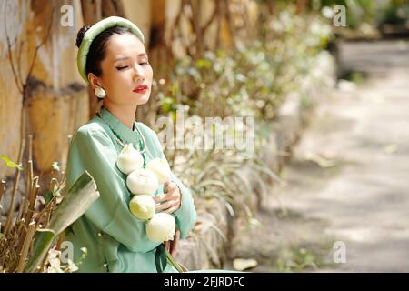 Pretty young Asian woman standing in park with bunch of lotus flowers, closing eyes and enjoying warm spring sunlight Stock Photo