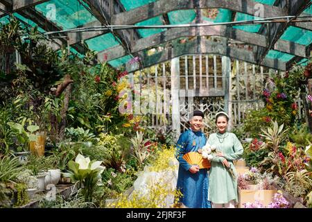 Happy young Vietnamese couple having wedding ceremony in greenhouse with many blooming flowers Stock Photo