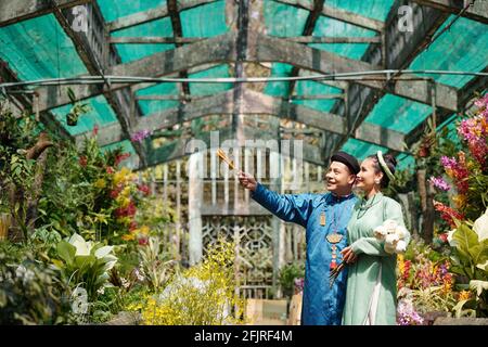 Happy young Vietnamese man showing beautiful flowers in greenhouse to bride Stock Photo