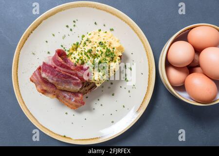 Scrambled eggs with bacon and chives on sourdough toast on plate with bowl of eggs to one side. Top down shot. Stock Photo