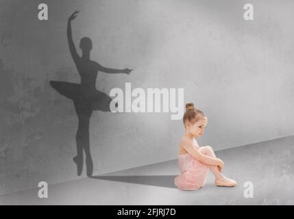 Cute little girl sitting on floor in dance studio, shadow of ballet dancer on grey wall, dreaming to become dancing star Stock Photo