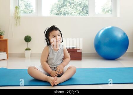 Adorable smiling little mixed-race kid sitting on yoga mat and listening to music in headphones Stock Photo