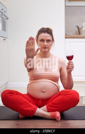 Fitness during pregnancy and abstinence from drinking wine. A pregnant woman at a sports training session at home shows a stop gesture as a sign of gi Stock Photo