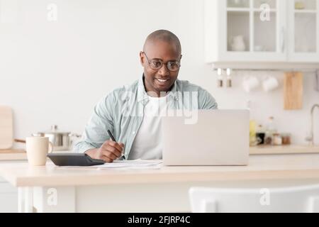 Plan finances. Positive african american mature man calculating taxes and using laptop computer, sitting in kitchen Stock Photo