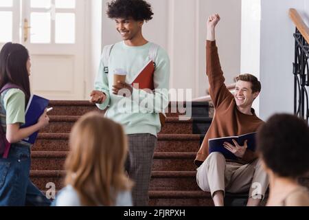 Cheerful student showing yes gesture near multiethnic friends on stairs on university Stock Photo