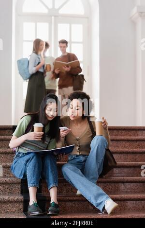 Interracial students with coffee to go, notebook and smartphone sitting on stairs Stock Photo