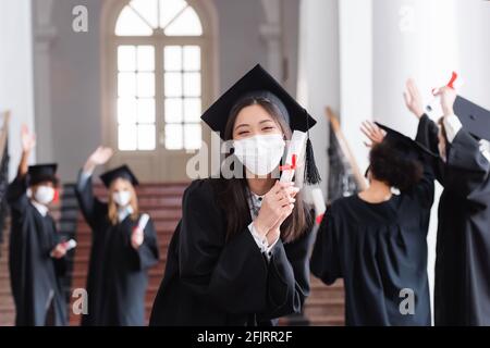 Asian bachelor in cap and medical mask holding diploma near blurred friends Stock Photo