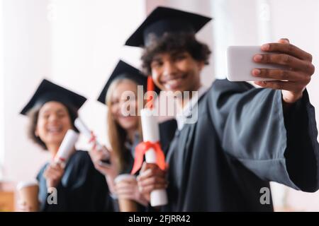 Blurred african american graduate taking selfie with smartphone near friends Stock Photo