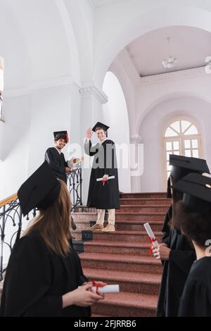 Smiling multiethnic students with diplomas waving at friends on blurred foreground Stock Photo