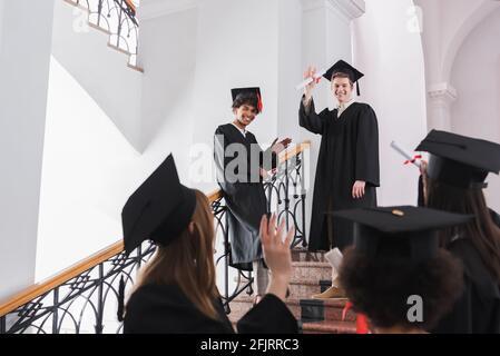Smiling multiethnic graduates with diplomas waving at friends on blurred foreground Stock Photo