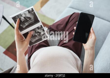 High angle view of pregnant woman sitting on sofa with x-ray images of her baby and using mobile phone Stock Photo