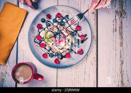 top view of a person's hands with knife and fork to eat a delicious breakfast of pancakes with fresh fruits and chocolate syrup and a hot coffee. Stock Photo