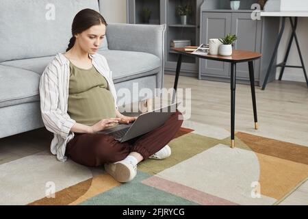 Young pregnant woman sitting on the floor with laptop on his knees and working online in the room at home Stock Photo