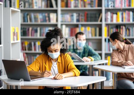 Students during pandemic with medical masks. Concentrated african american girl wears protective medical mask sit at table in library, preparing for exam, takes notes, classmates sit at background Stock Photo