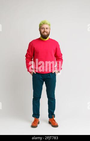 Full length photo of happy smiling bearded man with bright appearance, wearing red sweater, pants and shoes, keeps hands in pockets. Indoor studio sho Stock Photo