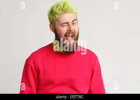 Cheerful playful young Caucasian man with beard in good mood, smiling broadly and winking at camera, keeps mouth open, dresses red jumper. Indoor stud Stock Photo