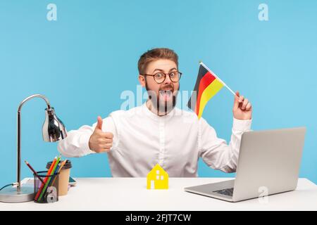 Positive excited man in eyeglasses showing thumbs up holding flag of germany, sitting workplace with paper house, satisfied with accommodation purchas Stock Photo
