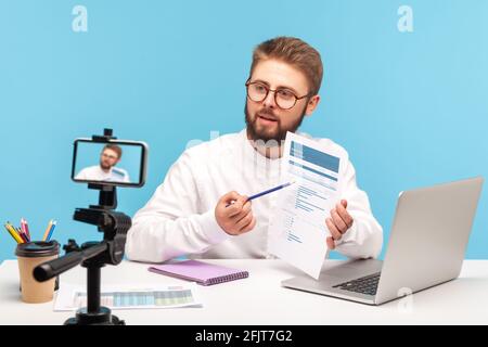 Concentrated bearded man analyst e[plaining how to work with datas sitting at workplace at office and posing at smartphone camera, recording tutorial. Stock Photo