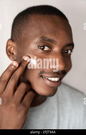Smiling young African man applying moisturizing cream to his cheek Stock Photo